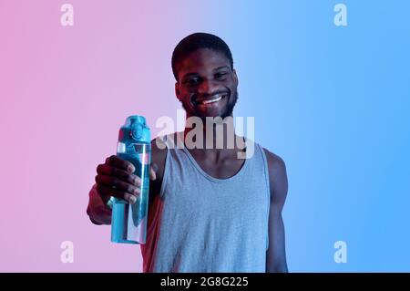 Lächelnder junger schwarzer Sportler zeigt Flasche mit frischem Wasser in Neonlicht. Fitness, aktiver Lebensstil, Wellness-Konzept Stockfoto