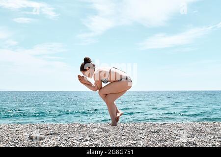 Schlanke Frau, die Garudasana im Stehen macht, Adler posieren am Strand mit blauem Meer im Hintergrund. Stärkung der Muskeln der Beine und Koordination des Körpers. Pra Stockfoto