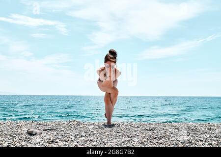 Schlanke Frau, die Garudasana im Stehen macht, Adler posieren am Strand mit blauem Meer im Hintergrund. Stärkung der Muskeln der Beine und Koordination des Körpers. Pra Stockfoto