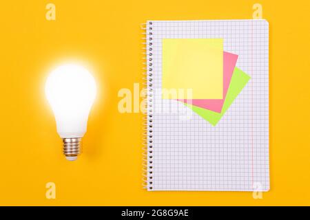 Ein Ideenkonzept - leuchtende Energiesparlampe und EIN Notizblatt mit Haftnotizen auf gelbem Tisch. Draufsicht, Flach Liegend, Leere Vorlage Stockfoto
