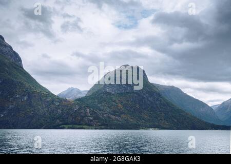 Majestätische Berge ragen an einem bewölkten Tag im arktischen Sommer über den See in Nordnorwegen. Kleine Häuser zwischen den Bergen weit in der Ferne Stockfoto