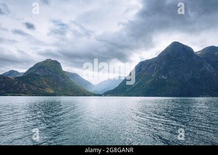 Majestätische Berge ragen an einem bewölkten Tag im arktischen Sommer über den See in Nordnorwegen. Kleine Häuser zwischen den Bergen weit in der Ferne Stockfoto