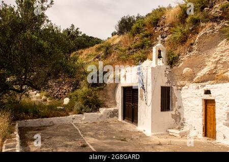 Alte weißgetünchte kleine christliche Kirche in einem Felsen auf einem Hügel auf der Insel Milos in der Nähe von Trypiti Stadt mit grünen Pflanzen in der Umgebung, Kykladen Inseln, Griechenland gebaut. Stockfoto