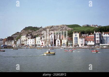 1975, ein Blick über den See mit Bootstouren zur Altstadt von Hastings an der Ostküste von Sussex, England, Großbritannien. Im Zusammenhang mit der berühmten Schlacht von Hastings im Jahr 1066, wurde es später zu einem der mittelalterlichen Cinque Ports. Mit der Entwicklung der Eisenbahn wurde sie im 19. Jahrhundert zu einem beliebten Badeort, und obwohl der Tourismus seine wichtigste Wirtschaft ist, war die Fischereiindustrie immer von Bedeutung. Die Stadt verfügt über die größte Strandfischerflotte Großbritanniens, die sich am Stade, einem Kiesstrand in der Altstadt, befindet. Stockfoto