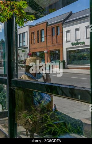 Dorking, Surrey, UK, 07-19-2021: Eine unbekannte Person, die mit Strohhut und Gesichtsmaske sitzt und an einer Bushaltestelle vor der lokalen Waitrose einen Zigarren rauchet. Stockfoto