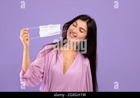 Coronavirus ist vorbei. Glückliche armenische Frau, die ihre Gesichtsmaske auf purpurem Studiohintergrund auszieht Stockfoto