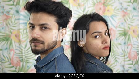 Junges indisches Paar, das mit ernstem Gesichtsausdruck auf dem Hintergrund eines Wals Rücken an Rücken steht Stockfoto