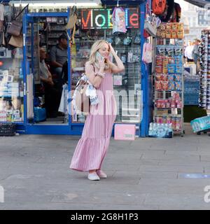 London, Greater London, England, Juni 12 2021: Junge Frauen in einem rosa Kleid mit einer braunen Handtasche am Telefon vor einem blauen Geschäft. Stockfoto