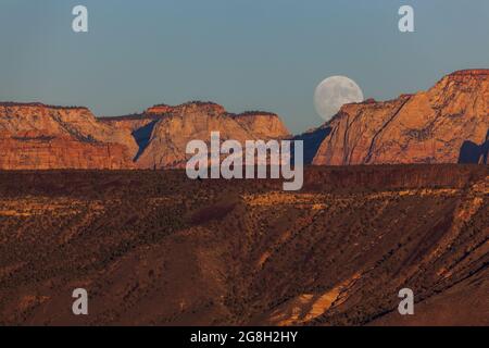 Der Vollmond, der über den dramatischen Bergen des Zion National Park aufgeht und von der untergehenden Sonne mit klarem Himmel erleuchtet wird. Stockfoto