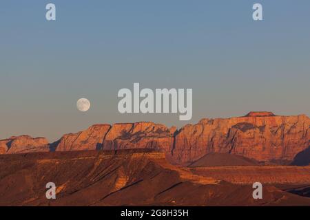 Der Vollmond, der über den dramatischen Bergen des Zion National Park aufgeht und von der untergehenden Sonne mit klarem Himmel erleuchtet wird. Stockfoto