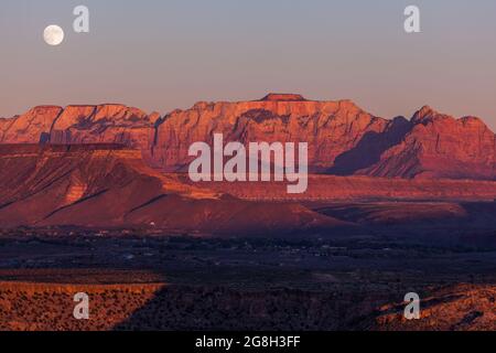 Der Vollmond, der über den dramatischen Bergen des Zion National Park aufgeht und von der untergehenden Sonne mit klarem Himmel erleuchtet wird. Stockfoto