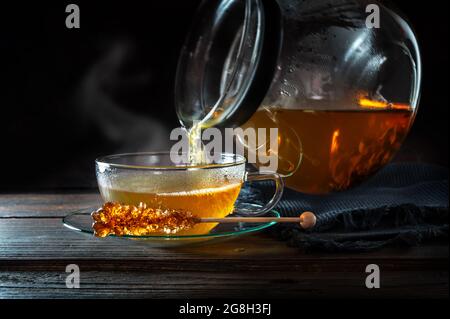 Heiß dampfender Tee wird aus einer Kanne in eine Glasschale gegossen, serviert mit einem Stab von Rock Candy, Bewegungsunschärfe, dunklem rustikalem Holzhintergrund mit Kopierfläche, Stockfoto