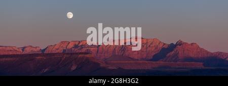Der Vollmond, der über den dramatischen Bergen des Zion National Park aufgeht und von der untergehenden Sonne mit klarem Himmel erleuchtet wird. Stockfoto