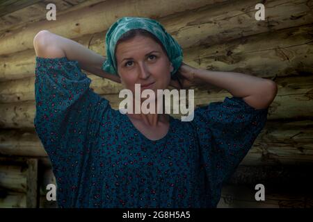 Frau in traditionellem Bauernkleid bindet ein Kopftuch vor dem Hintergrund einer Blockwand Stockfoto