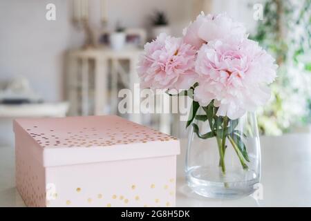 Frisches Bouquet von zarten rosa Pfingstrosen Blumen in Vase und Geschenkbox auf weißem Tisch mit hellen klassischen Innenarchitektur Hintergrund. Festliche Blumen für Stockfoto