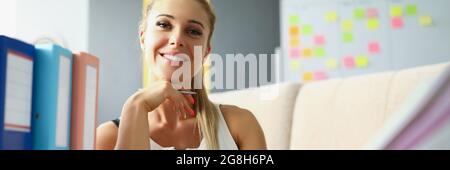 Glückliche blonde Frau sitzt am Tisch im Büro und lächelt. Am Arbeitsplatz befinden sich Ordner mit Dokumenten Stockfoto