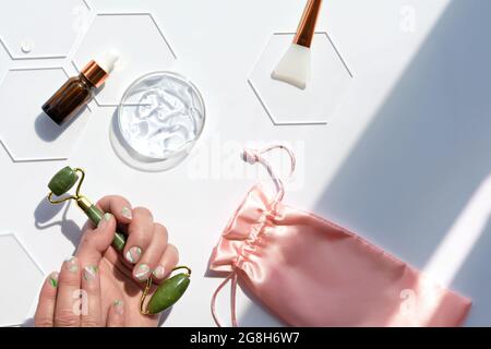 Hand mit Jade Gesichtswalze und Feuchtigkeitsgel in Runde Glasschale. Hausgemachte Beauty-Gesichtsmassage-Therapie. Off weiß flach legen mit Petrischale und Stockfoto