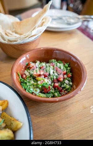 Nahaufnahme eines leckeren Tabbouleh-Salats mit Granatapfelkernen in einer braunen Keramikschale, Teller im Hintergrund auf einem Holztisch Stockfoto