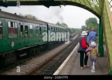 Dampfzug auf dem Bahnsteig des Bahnhofs von Corfe Castle mit Reisenden, die den Bahnsteig entlang laufen. Corfe Castle, Dorset - England. Stockfoto