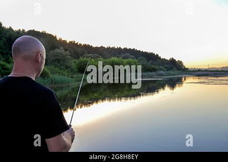 Ein Mann steht mit dem Rücken und fängt einen Fisch mit einer Angelrute im See, Sportfischen. Stockfoto