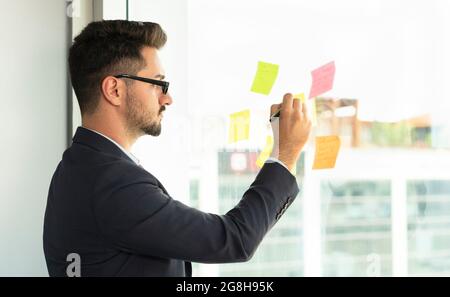 Porträt einer konzentrierten männlichen Führungskraft mit Brillen, die auf Haftnotizen an der Glaswand im Büro schreiben. Geschäftsmann, der sich auf geschäftliche Aktivitäten konzentriert Stockfoto