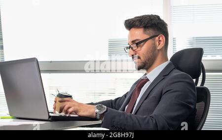 Porträt eines glücklichen Geschäftsmannes in formeller Kleidung, der im Büro mit einem Laptop arbeitet. Fröhlicher, entspannter Mann im Anzug, der beim Kaffee- oder Teetrinken arbeitet. Stockfoto