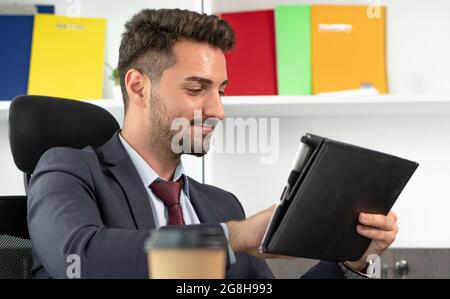 Fröhlicher junger professioneller Geschäftsmann mit digitalem Tablet, der im modernen Büro am Schreibtisch sitzt. Stockfoto