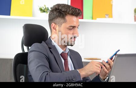 Junger professioneller Geschäftsmann in formeller Kleidung mit Smartphone, während er im modernen Büro am Schreibtisch sitzt. Stockfoto