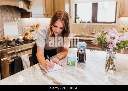 Frau schreibt im Planer mit Kaffee und Blumen in ihrer Küche Stockfoto