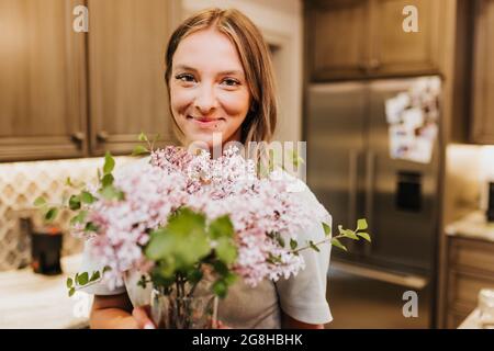 Frau lächelt die Kamera an, während sie eine Vase voller Flieder hält Stockfoto