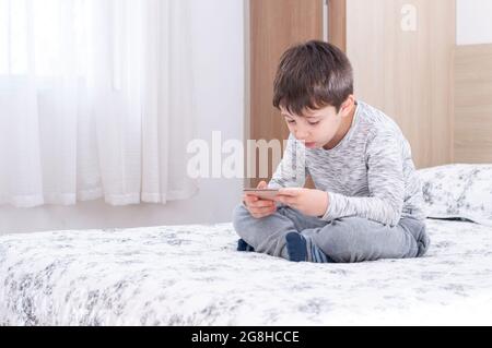 Kleiner 6 Jahre alter Junge, der Smartphone benutzt und spielt. Netter kleiner Junge, der im Bett Spiele auf dem Smartphone spielt. Boy spielt in Videospiel auf seinem Smartphone. Stockfoto