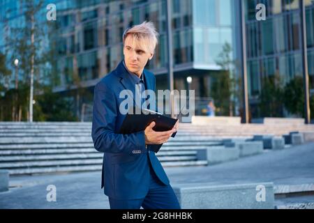 Vor einem Bürogebäude steht ein junger Mann im Geschäftsanzug mit einem Papierordner in der Hand. Stockfoto