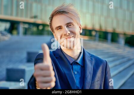 Fröhlicher Geschäftsmann in Anzug zeigt Daumen in der Nähe der Treppe des Bürozentrums. Stockfoto