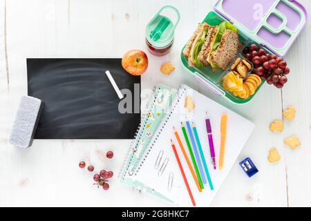 Draufsicht auf eine Auswahl an Schulmaterial und Mittagessen sowie eine leere Tafel, Kreide und Radiergummi. Stockfoto