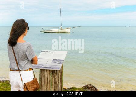 Eine alleinreisende Frau im Vordergrund liest eine Informationstafel über den Perlenfeger, der im Hintergrund in Shark Bay Western Australia verankert ist. Stockfoto