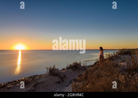 Eine alleinreisende Frau steht auf einer Sanddüne und bewundert den klaren, goldenen Sonnenuntergang an der Küste des Ningaloo an der Coral Bay in Westaustralien. Stockfoto