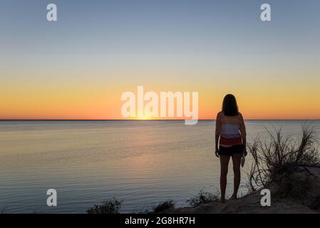 Eine alleinreisende Frau steht auf einer Sanddüne und bewundert den klaren, goldenen Sonnenuntergang an der Küste des Ningaloo an der Coral Bay in Westaustralien. Stockfoto