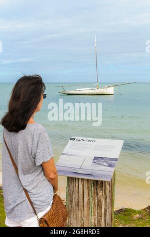 Eine alleinreisende Frau im Vordergrund liest eine Informationstafel über den Perlenfeger, der im Hintergrund in Shark Bay Western Australia verankert ist. Stockfoto