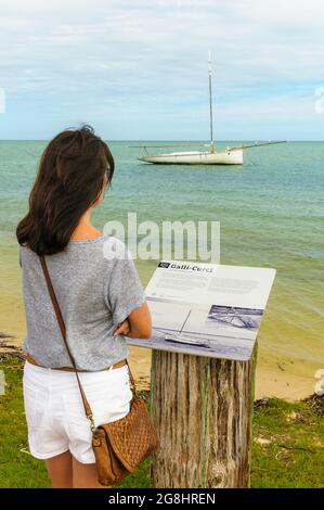 Eine alleinreisende Frau im Vordergrund liest eine Informationstafel über den Perlenfeger, der im Hintergrund in Shark Bay Western Australia verankert ist. Stockfoto