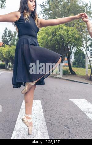 Nahaufnahme einer Balletttänzerin in einem schwarzen Kleid und Ballettschuhen, die die Hand einer anderen Person auf einem Fußgängerüberweg halten Stockfoto