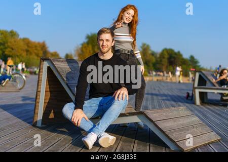 Glückliches Paar ein Mann und ein Mädchen mit langen roten Haaren sitzen in einer Umarmung auf einem Holzdeck. Junge Männer und Frauen kaukasischer Ethnie in legerer Kleidung an einem warmen sonnigen Tag umarmen Stockfoto