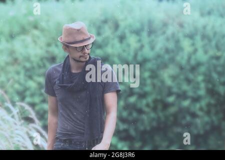 Attraktiver Mann, der im Regen hinter der Baumnatur herumläuft, im Retro-Stil, mit Hut, Brille und Schal. Stockfoto