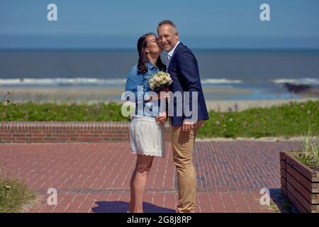 Fröhliches Hochzeitspaar, das die Kamera anschaut, während sie Hand in Hand auf einem Gehweg stehen, der einen tropischen Strand und das Meer überblickt, während sie ein römer sind Stockfoto