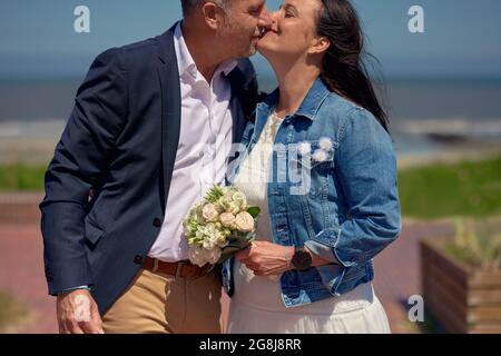 Fröhliches, romantisches, reifes, frisch verheiratetes Paar, das sich an ihrem Hochzeitstag auf einer Promenade vor dem Hintergrund des Ozeans in formeller Kleidung und Holdin küsst Stockfoto