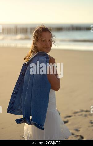 Attraktive Frau mittleren Alters in stilvollem weißen Kleid trägt einen Blumenstrauß und Jacke über ihre Schulter, die über einen einsamen Strand A läuft Stockfoto