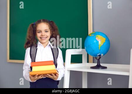 Lächelndes Mädchen mit Rucksack, der Bücherhaufen in der Nähe der Tafel im Klassenzimmer hält. Nette Schülerin, die gerne wieder zur Schule geht Stockfoto