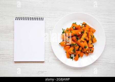 Hausgemachte geröstete Karotten mit Kräutern auf einem weißen Teller, leerer Notizblock auf weißem Holzhintergrund, Draufsicht. Stockfoto