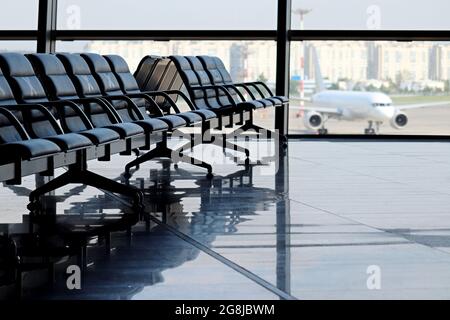 Leere Wartesessel im Flughafengebäude gegen das Flugzeug auf der Start- und Landebahn. Reisen während der Quarantäne bei einer Coronavirus-Pandemie Stockfoto