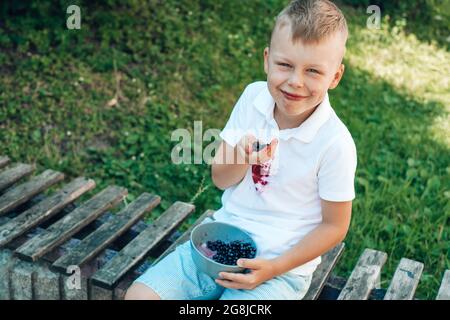 Hübscher Junge, der mit den Händen frische Beeren isst und auf einer Holzbank sitzt. Im Freien. Reinigungskonzept für Flecken Stockfoto