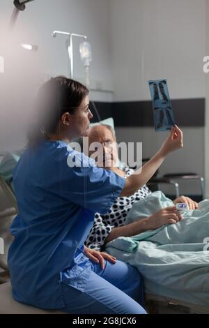 Alte Patientin mit Lungenerkrankung, die mit einer im Krankenhausbett liegenden Sauerstoffmaske atmet, lauschende Krankenschwester, die vor der Operation eine Röntgendiagnostik zeigt. Stockfoto